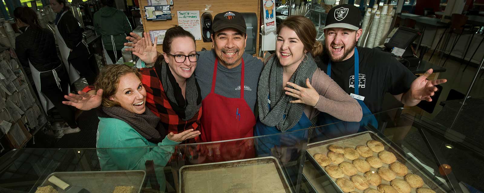 Three women and two men in a group hug behind trays of cookies