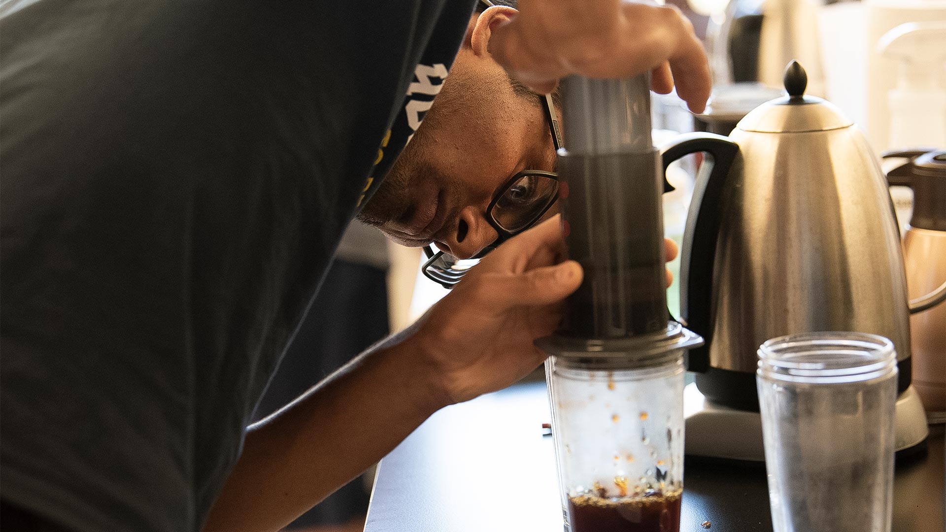 A coffee class student bends down to look carefully at a glass sitting under a filter with coffee in it as it drips into the glass.