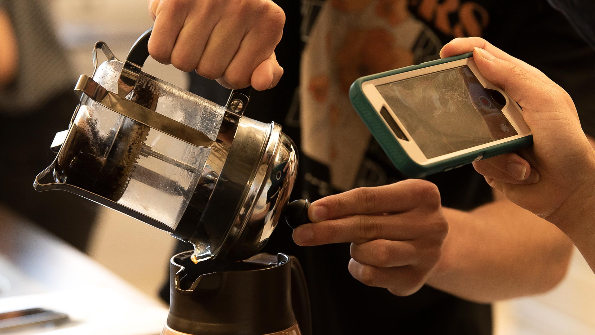 Closeup photo of a student's hands as coffee is being poured from a French press into a carafe. Another student's hands are shown holding a smartphone and taking video of the pour.