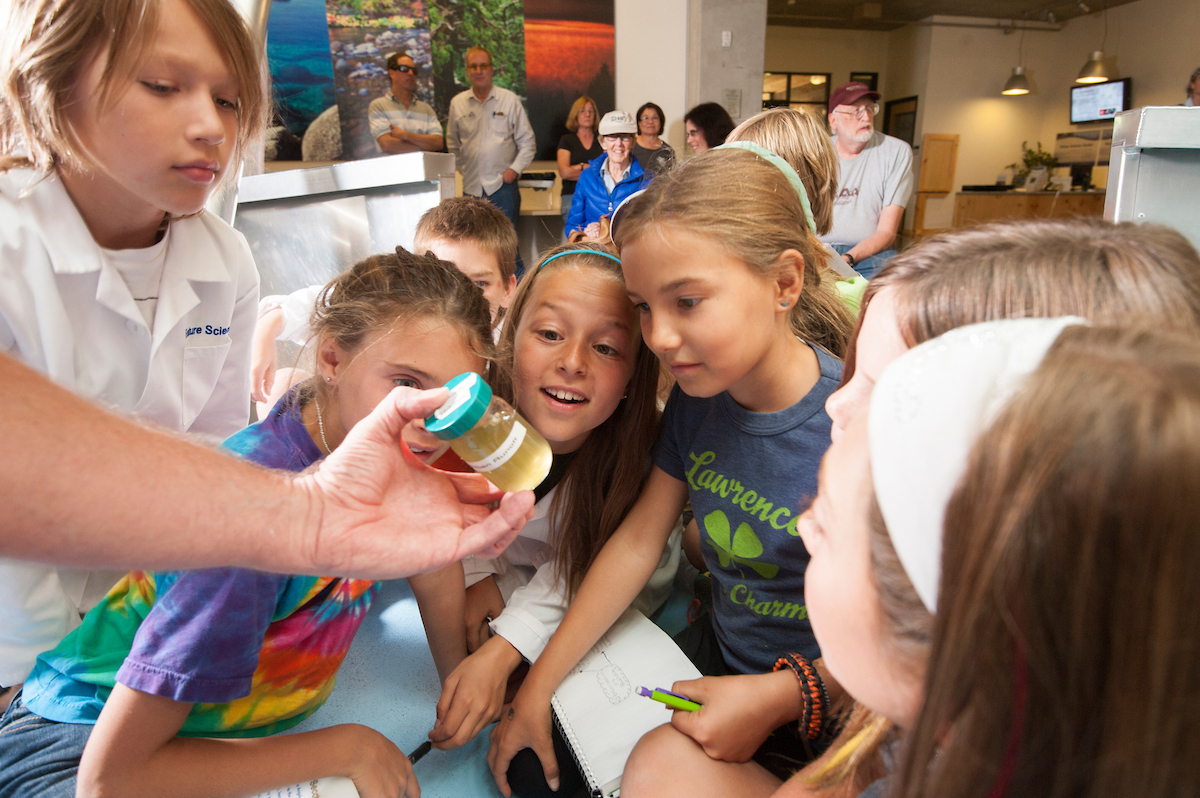 Excited children crowd around an adult holding a small jar of water, classroom photo