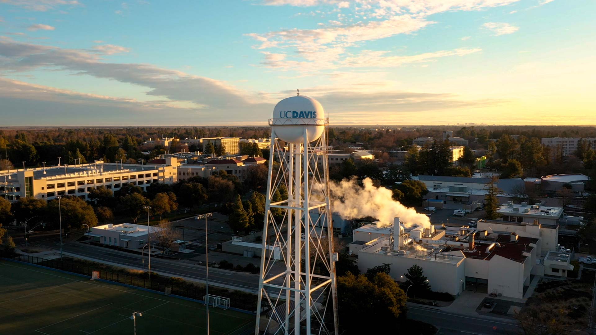 Aerial photo of a UC Davis water tower surrounded by campus. The sky is blue and gold at sunrise.