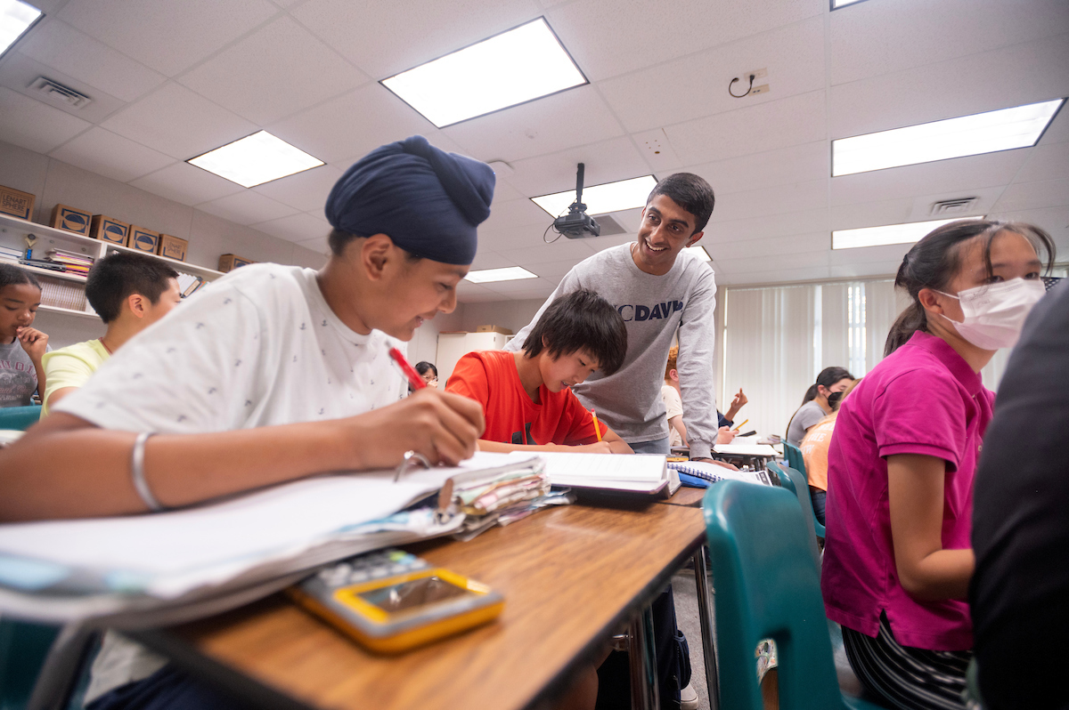 A college student leans down to help two high school students learn about spheres.