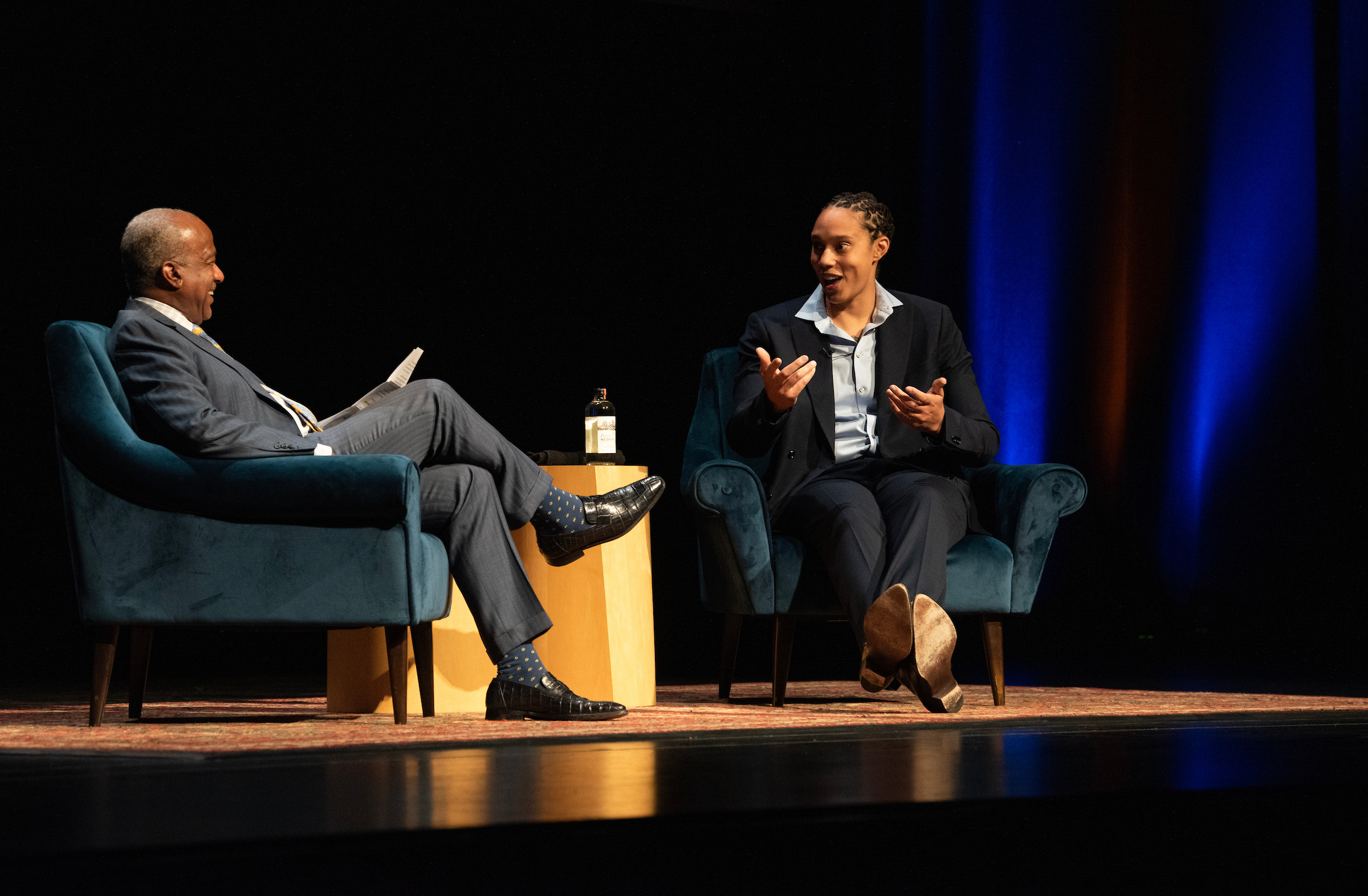 Two individual are seated in chairs on an illuminated theater stage. On the left in a light blue suit with their legs crossed in Chancellor Gary May. On the right is Brittney Griner, sitting in a dark blue suit, facing the Chancellor with her hands near her chest. 