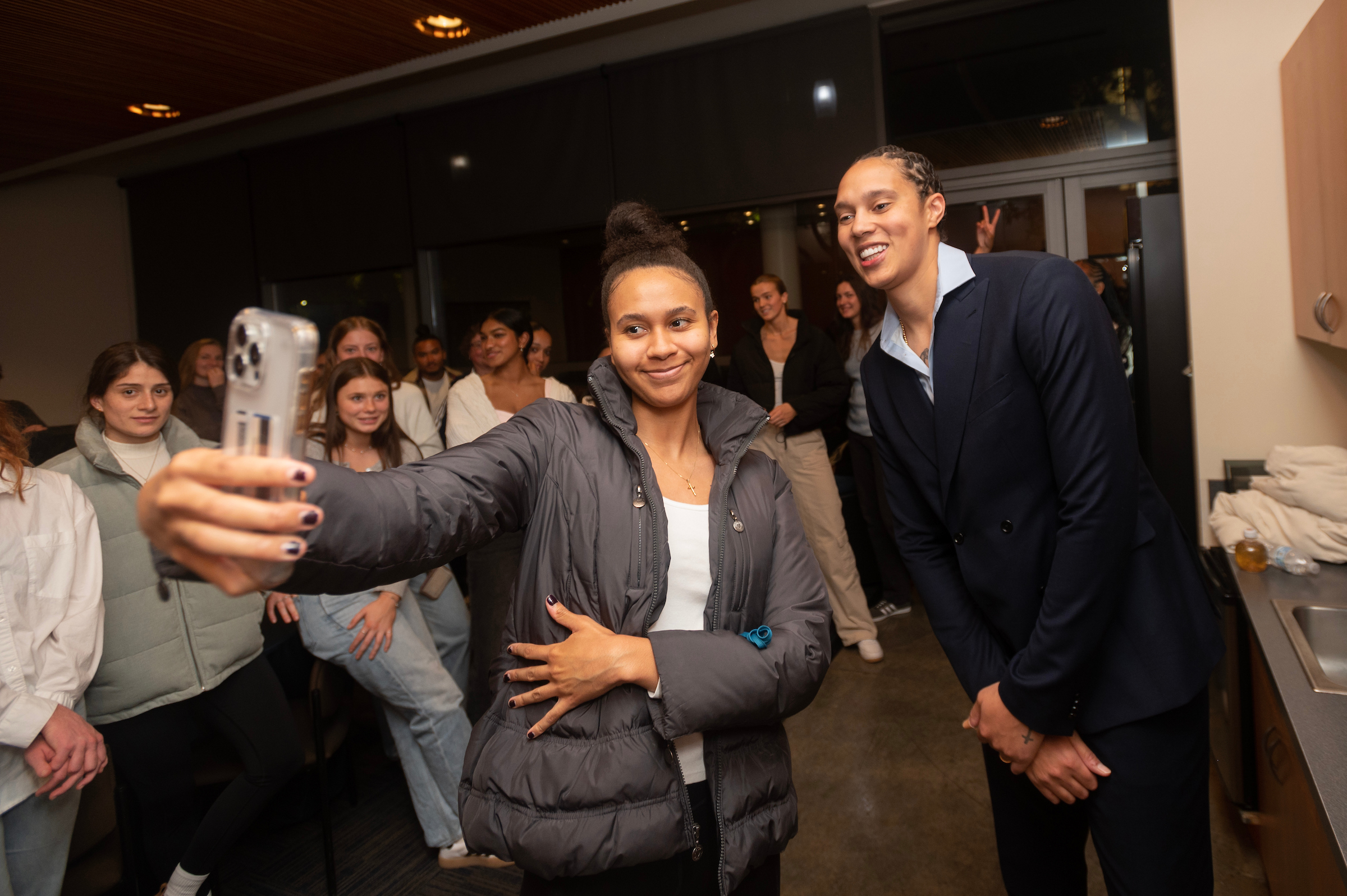 A young woman in a grey puffy coat is taking a selfie with Brittney Griner who is standing with her hands crossed in a dark blue suit. 