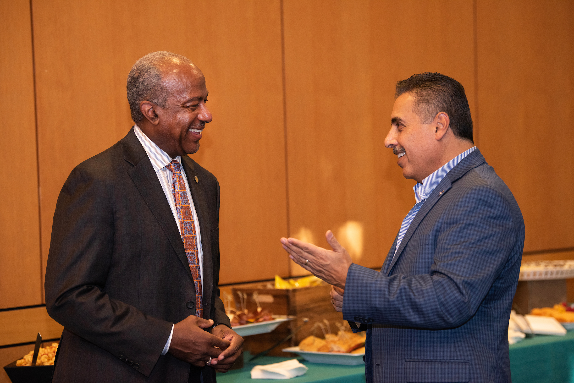 Two men smile in conversation with each other in front of a buffet setting.