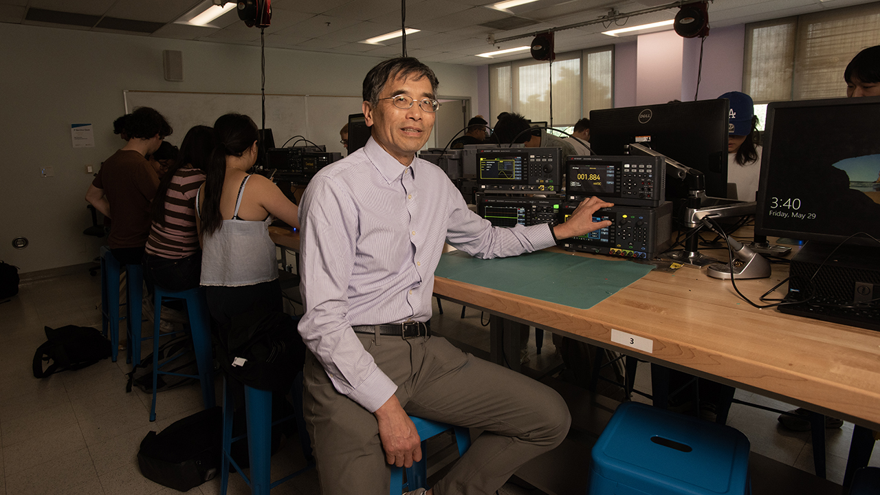Man seated at electronics workbench with students in background