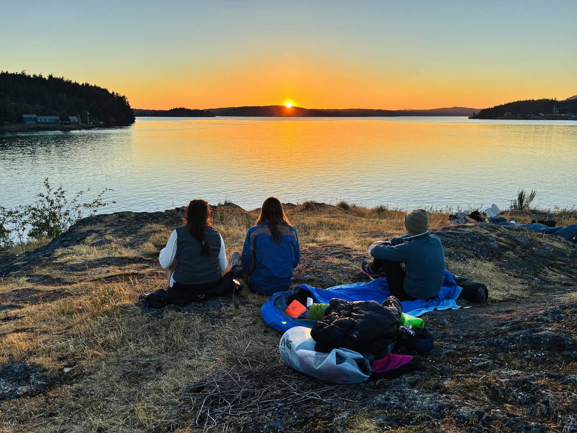 Three people sitting on rocky shoreline watching sunset over calm lake, bags in foreground