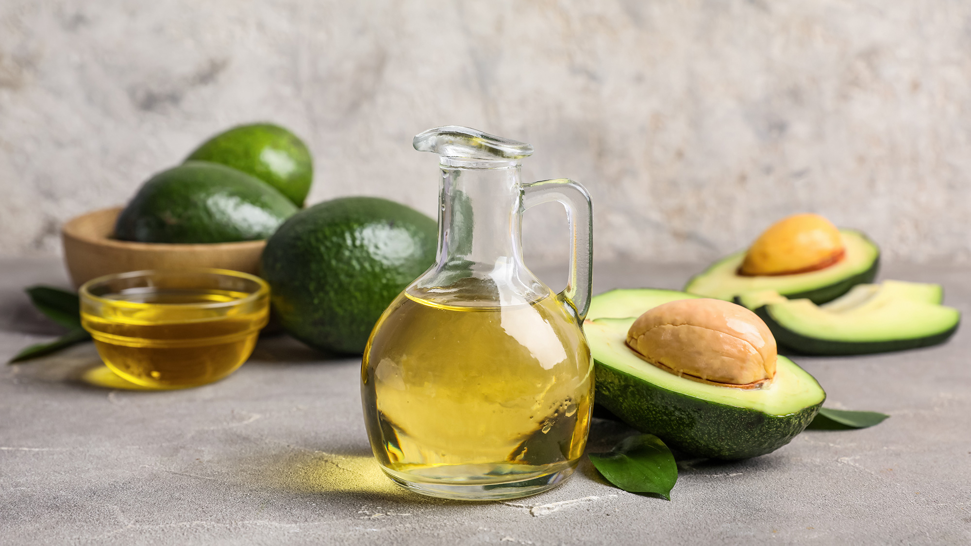 Glass bottle and bowl of golden avocado oil with whole and halved avocados on gray surface