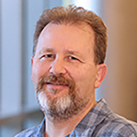 Face of a white man with brown hair and brown and grey facial hair, wearing a blue check shirt. 