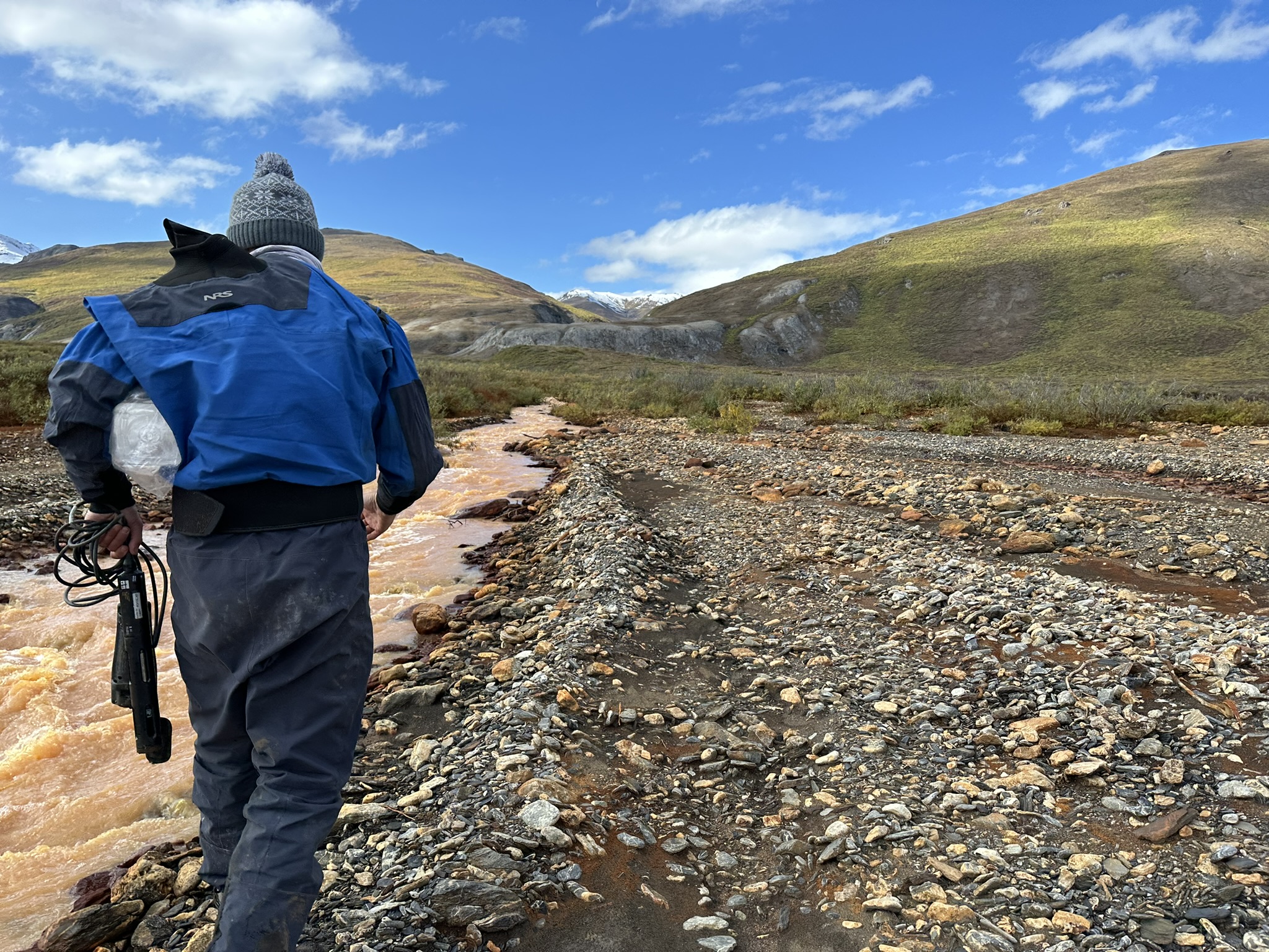 Scientist Brett Poulin in blue jacket carrying sampling equipment trots along a muddy tributary of a river under blue sky
