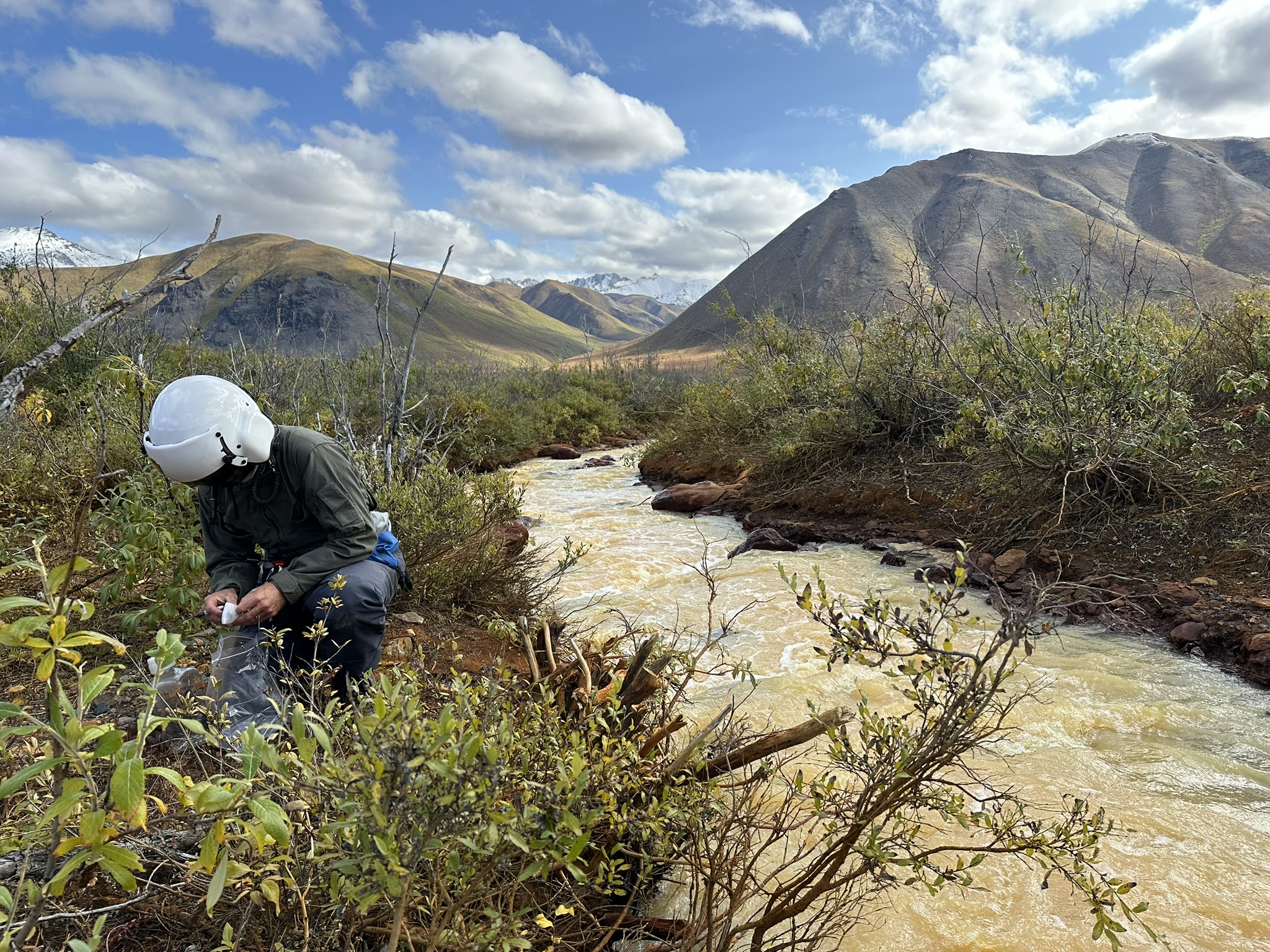 Scientist in protective white helmet crouching by muddy stream in Alaska, mountains under blue sky