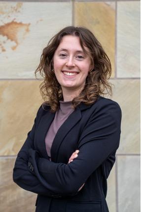 Colette Daulton smiles with her arms crossed in front of a beige stone wall.