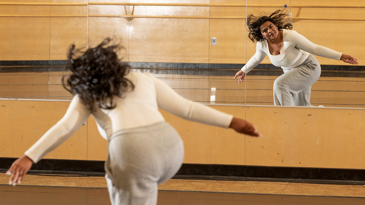 A woman in gray sweatpants and cream top dances facing a studio mirror.