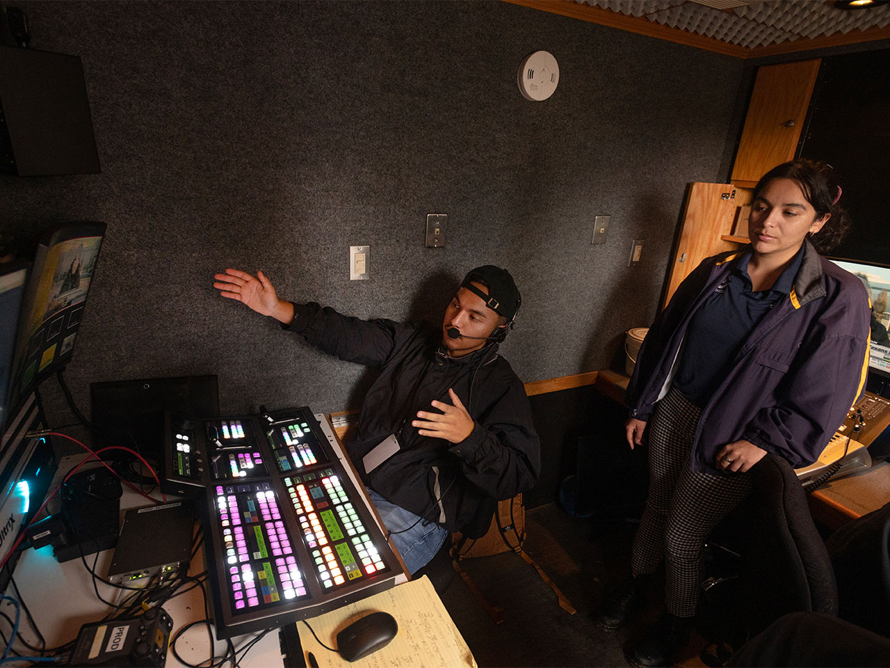 A director sits in front of an electronic switchboard and points to monitors while explaining film strategy to a UC Davis student worker.