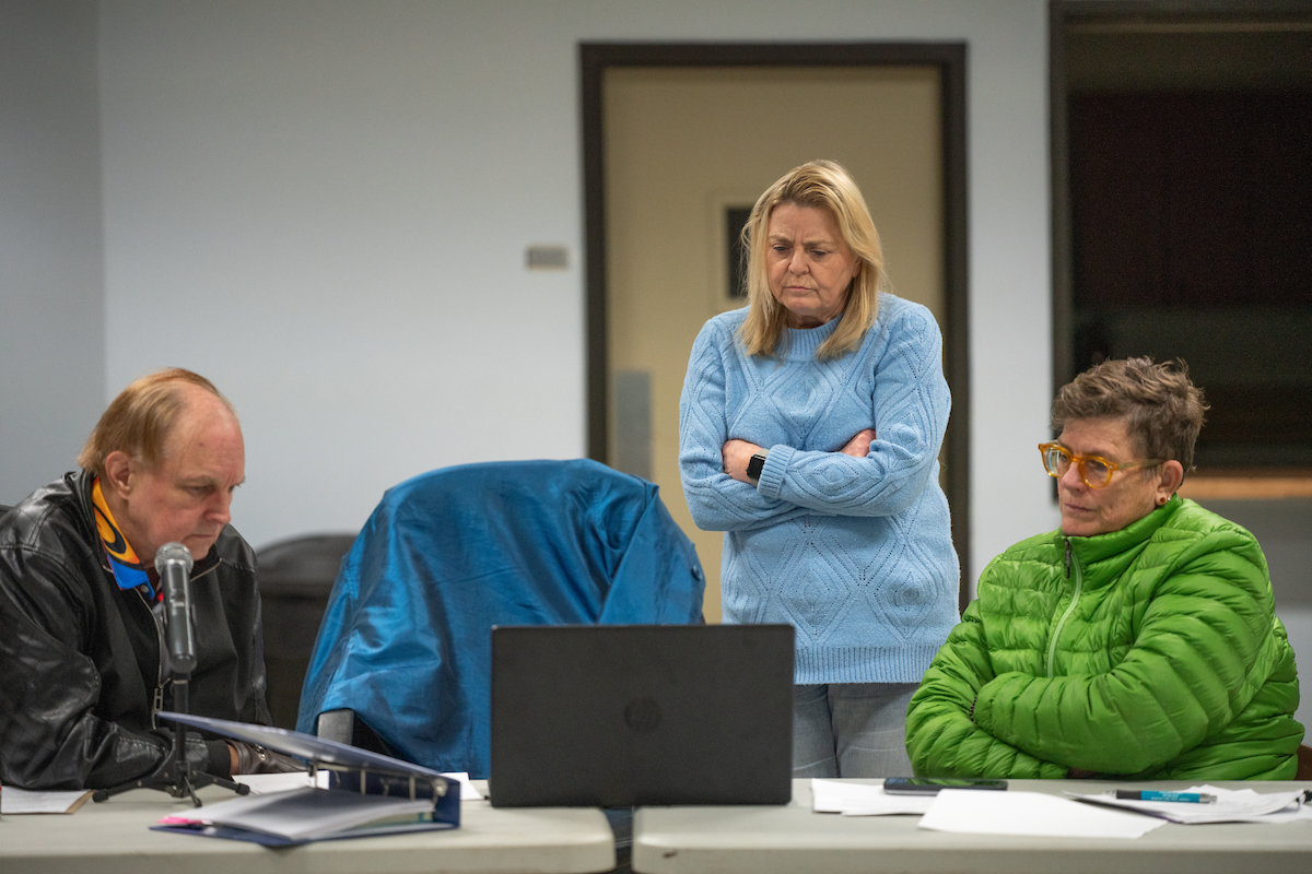 David Kent seated in black jacket, Kathleen Schaefer standing in blue coat with arms crossed and Iva Walton seated in green coat meet around a table.