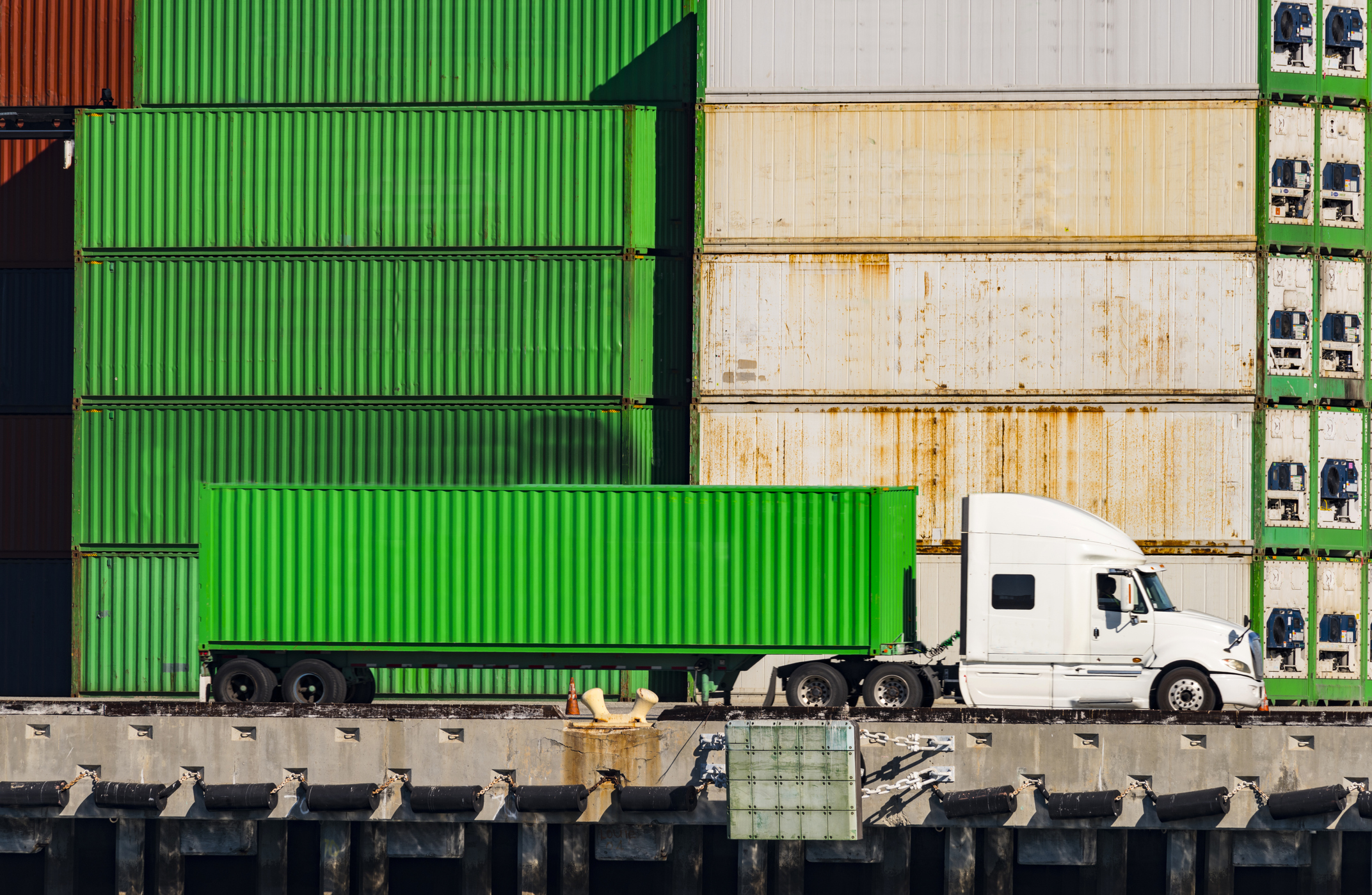 bright green heavy-duty truck sits parallel to stack of cargo containers at shipping port