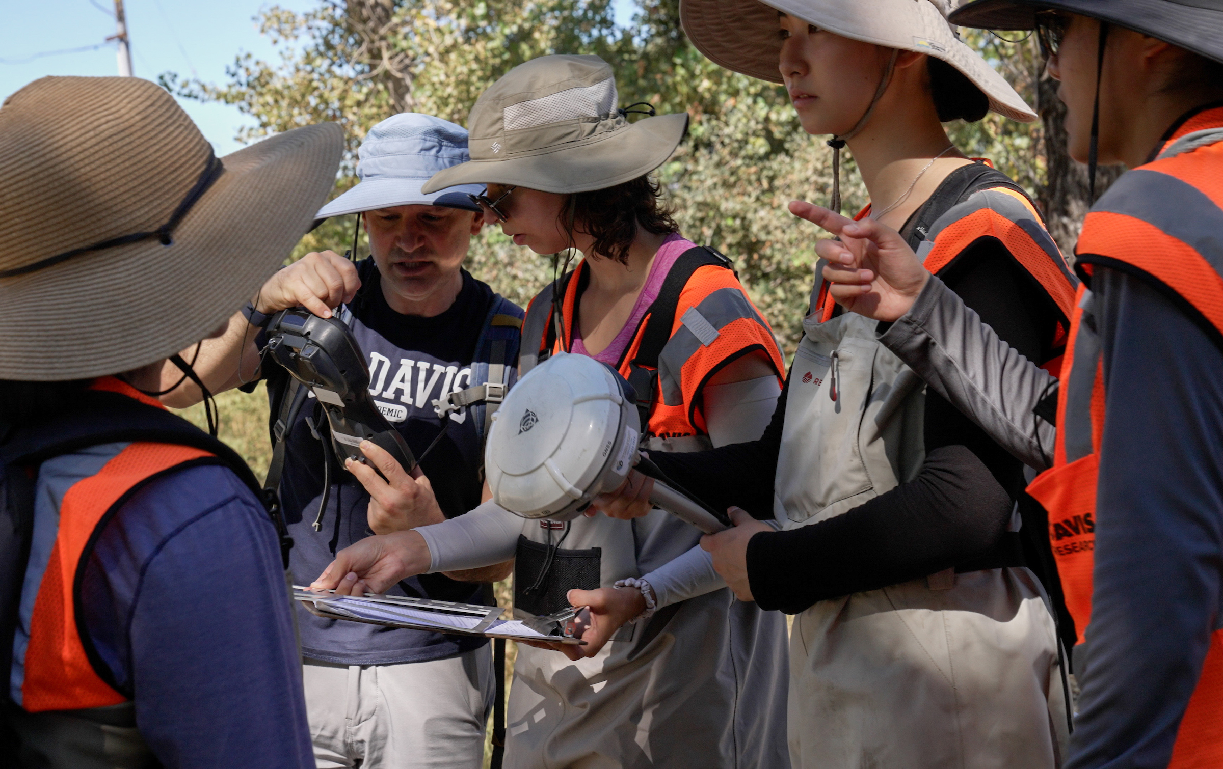 Researchers in hats and orange vests huddle around Gregory Pasternack in UC Davis t-shirt and hat outside looking at scientific device