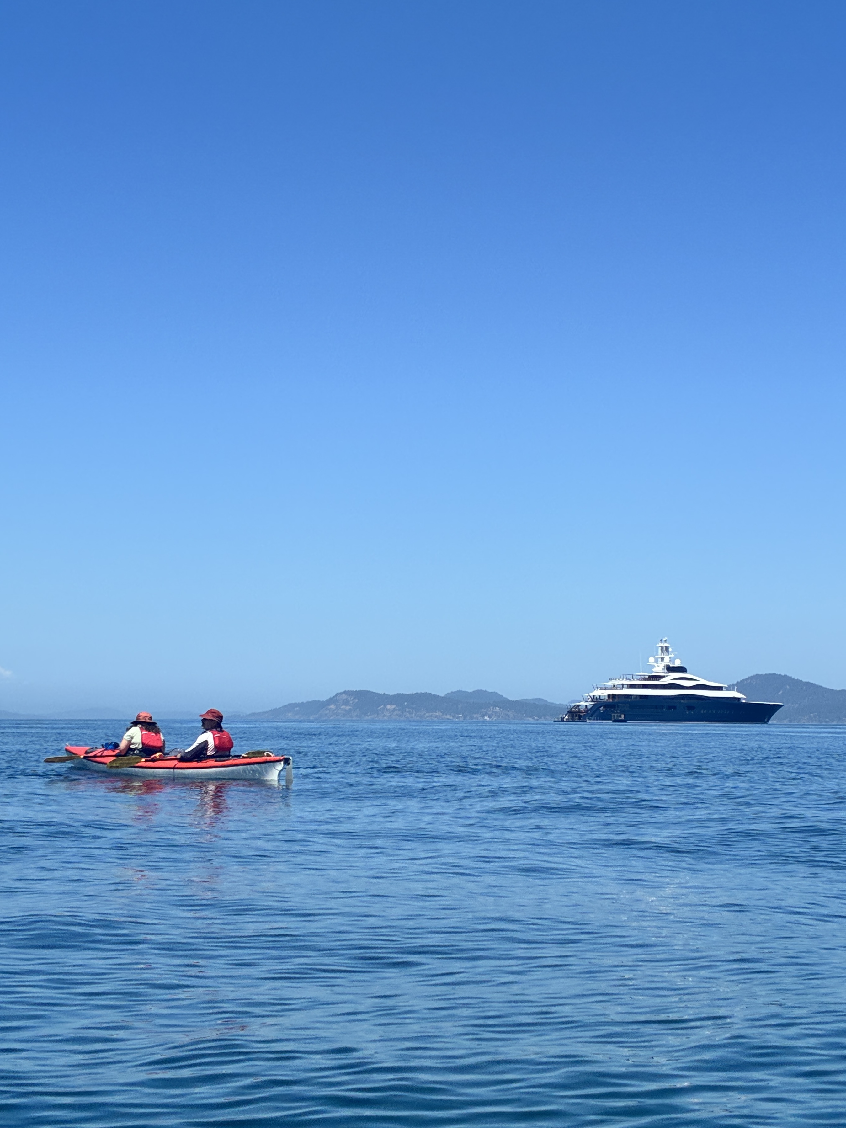 Two people kayaking in a red kayak near a distant white yacht on calm blue sea under clear sky
