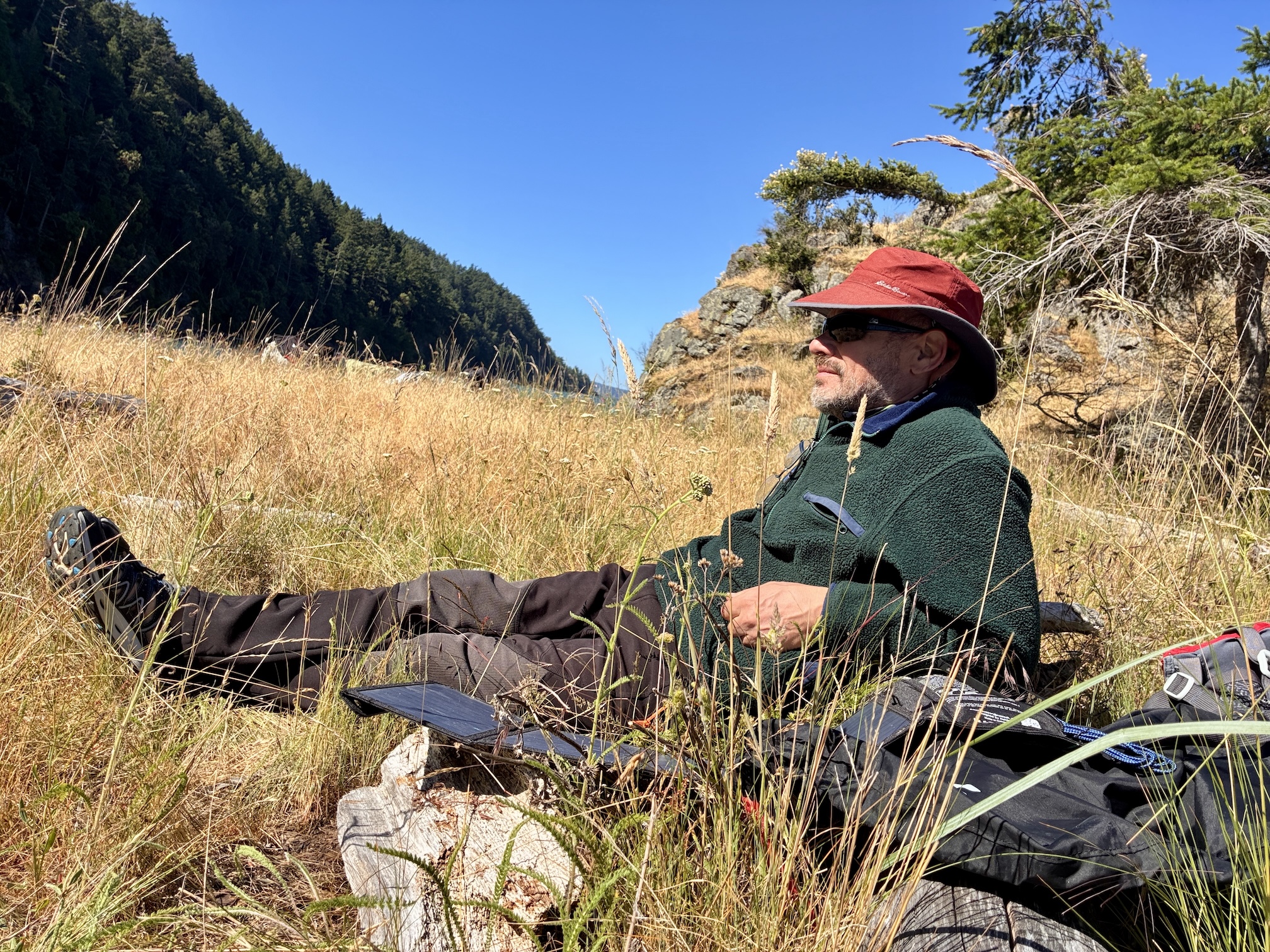 Nicholas Pinter reclining in grassy island meadow wearing red hat and green sweater beside backpack