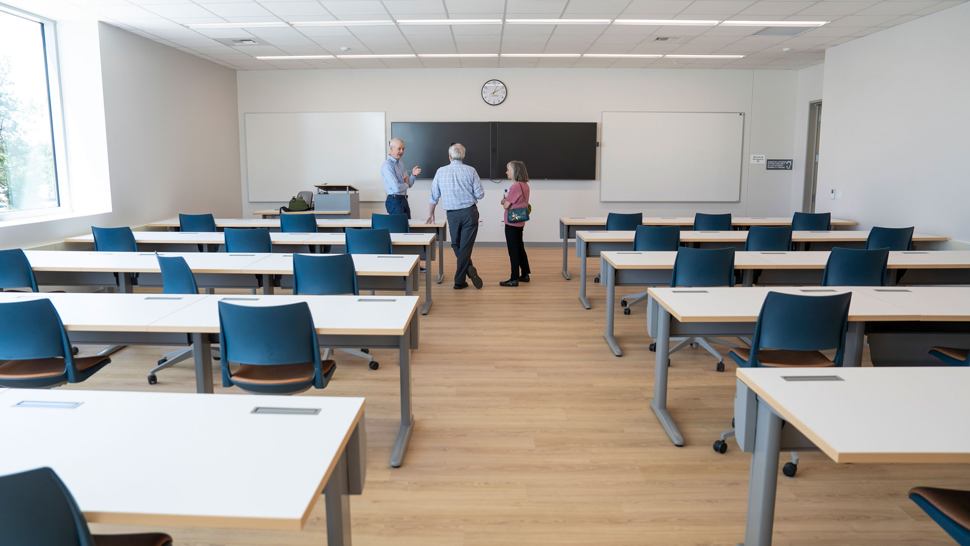Classroom with rows of white desks and blue chairs, three people talking at front