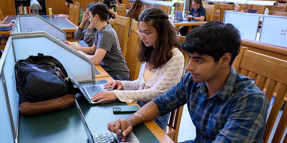Three students studying on laptops at a wooden library table