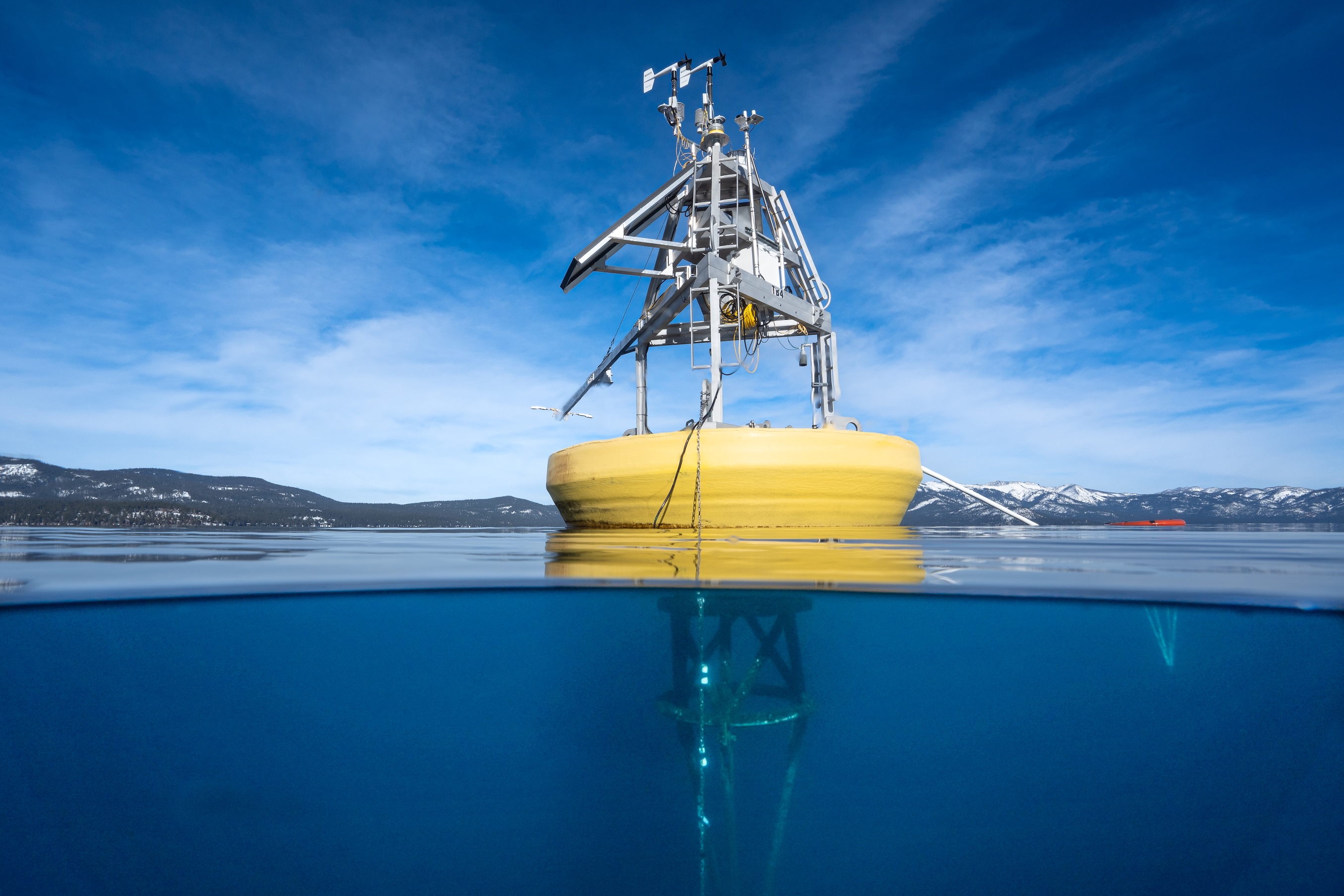 Yellow research buoy half above and below clear blue water, snowy mountains in background