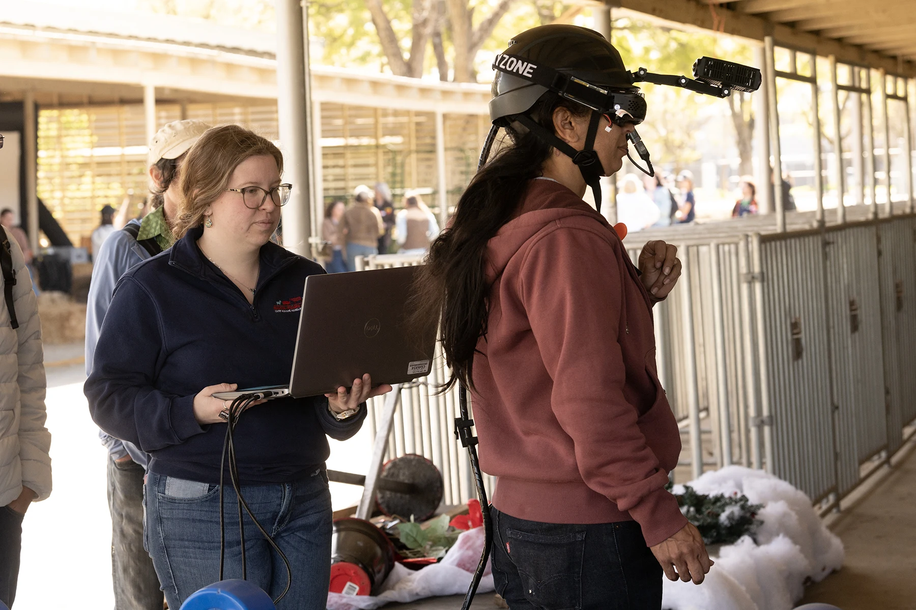 Two women in barn aisle, one wearing helmet-mounted camera, other holding laptop
