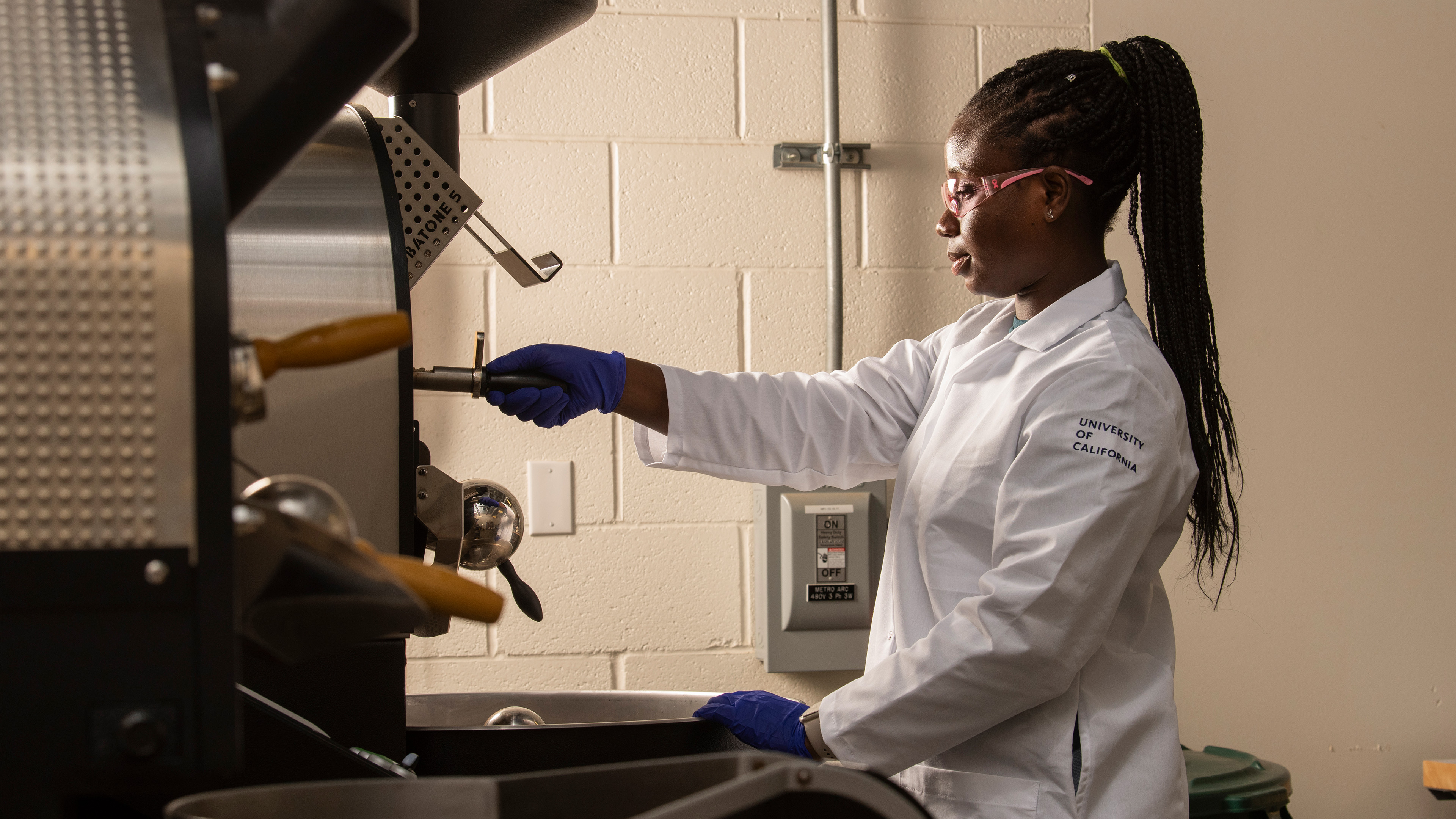 Laudia Anokye-Bempah, in full lab gear and a high ponytail, uses a stainless steel coffee roaster.