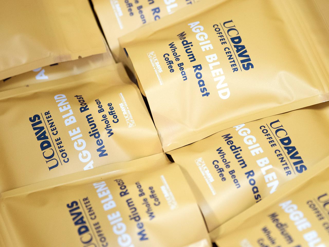 A closeup shot of many bags of coffee in a box. The bags are UC Davis branded in Aggie Gold with Aggie Blue text that says, "UC Davis Coffee Center. Aggie Blend. Medium Roast. Whole Bean Coffee."