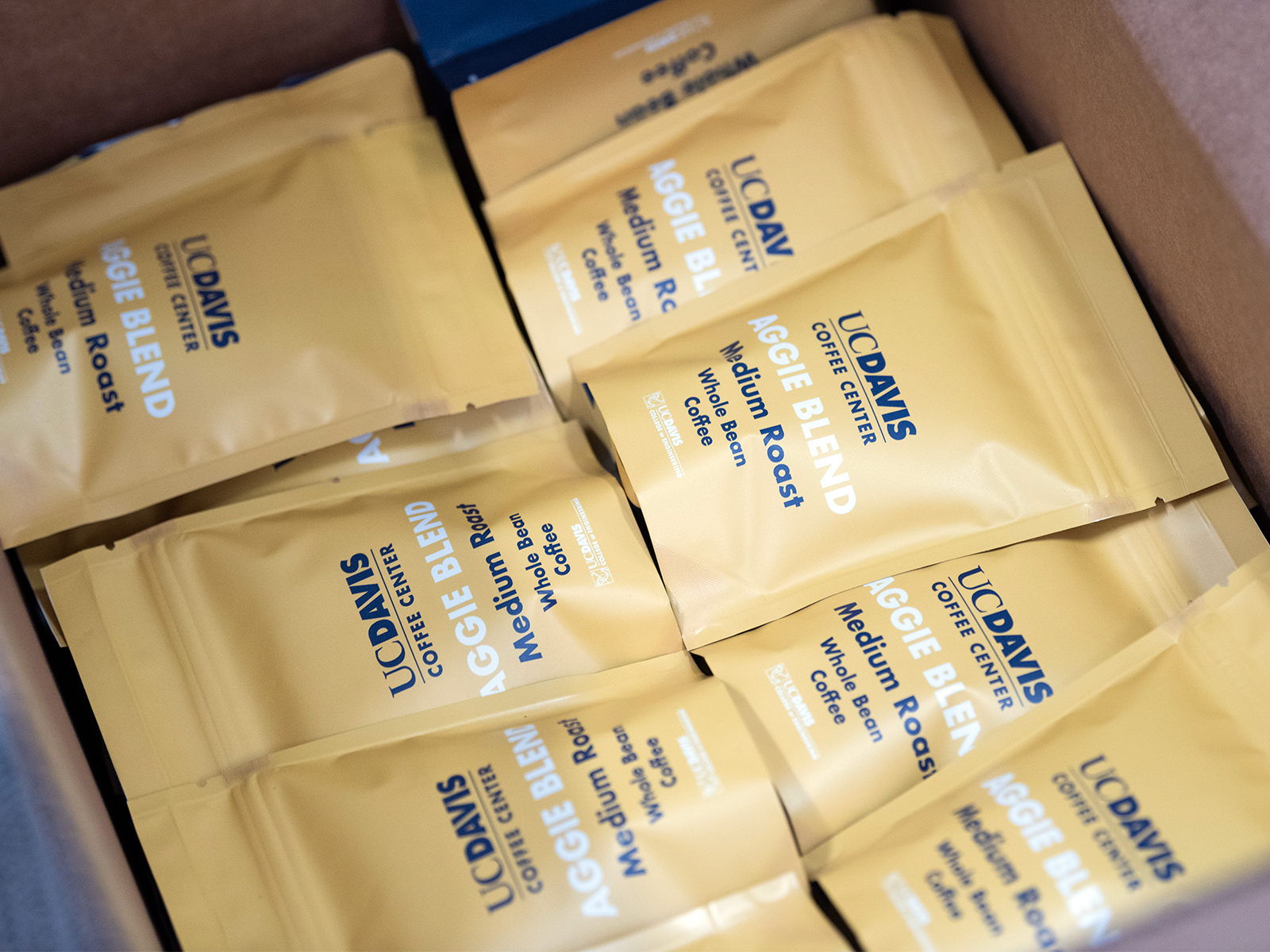 A closeup shot of many bags of coffee in a box. The bags are UC Davis branded in Aggie Gold with Aggie Blue text that says, "UC Davis Coffee Center. Aggie Blend. Medium Roast. Whole Bean Coffee."