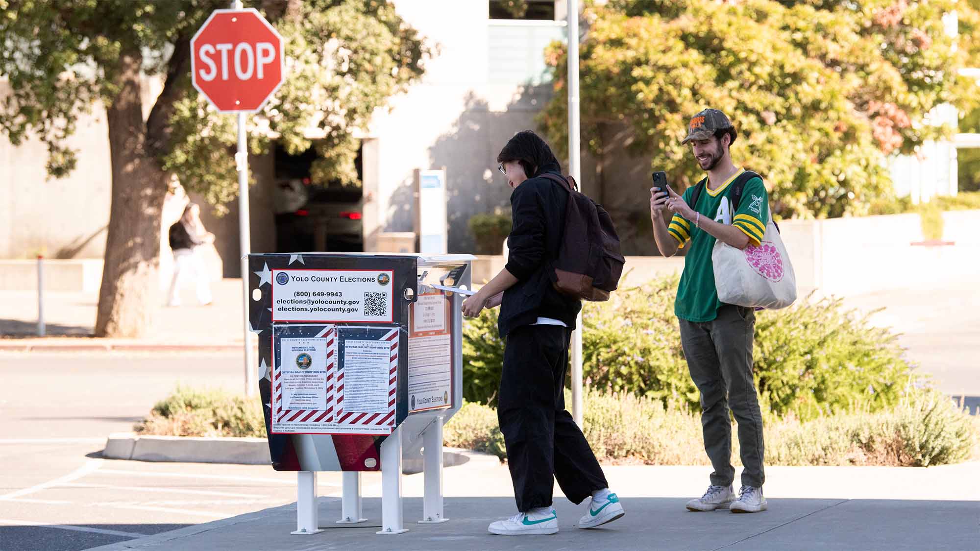 A person drops a ballot into a ballot box at UC Davis while another person takes a photo.