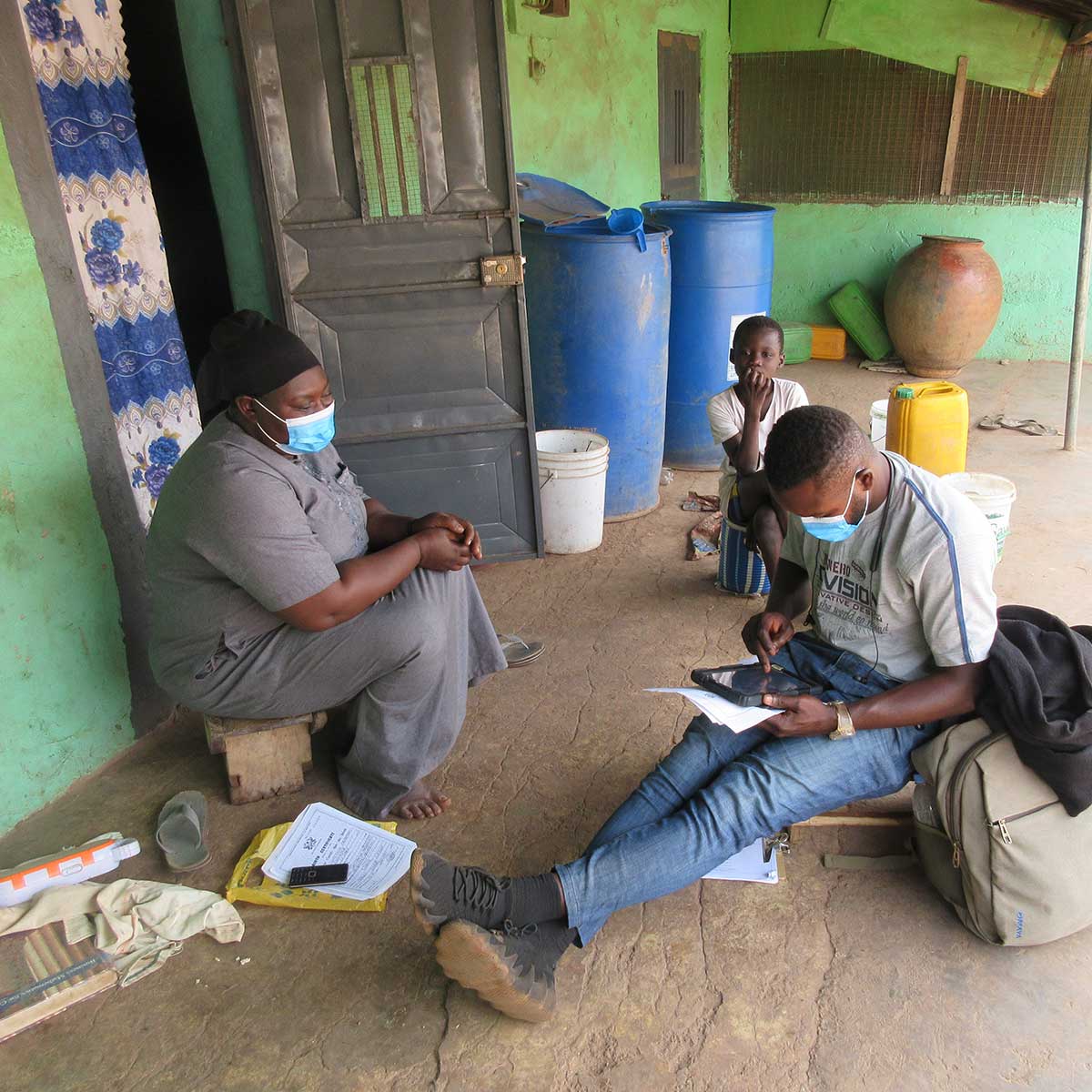 People sit on the ground outside the door of a house. One is recording something on a tablet while another talks.