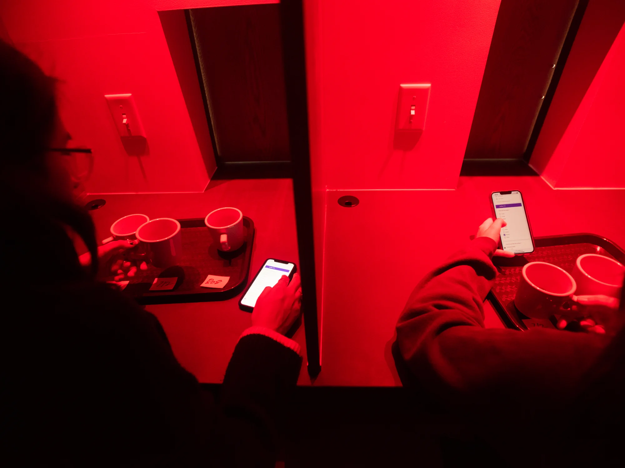 Two students sit in adjacent sensory booths, scrolling on their phones to check boxes beside different flavor descriptors. In front of each student is a tray holding three coffee mugs. In front of each mug is a three-digit, hand-written number on a piece of paper. The photo is lit only by red light.