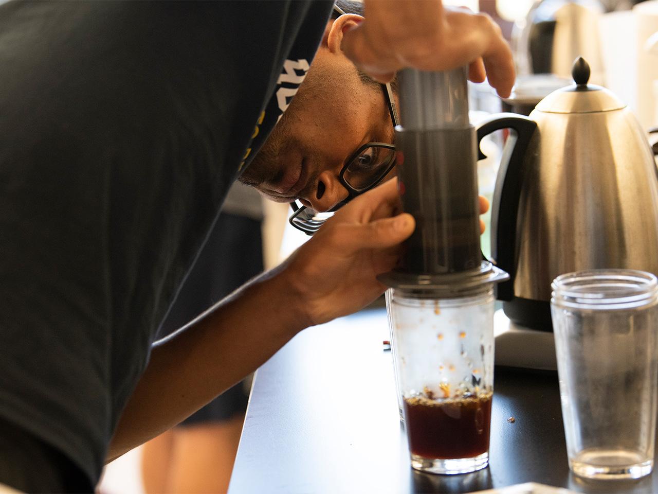 A student wearing a dark T-shirt and glasses leans over a lab table, which is filled with coffee and science instruments, to push hard on a device that drips coffee into a clear cup.