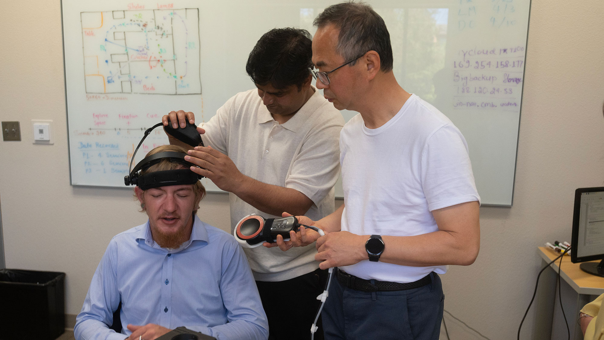 Three men in lab adjusting a wearable headset on a seated man, focused collaboration