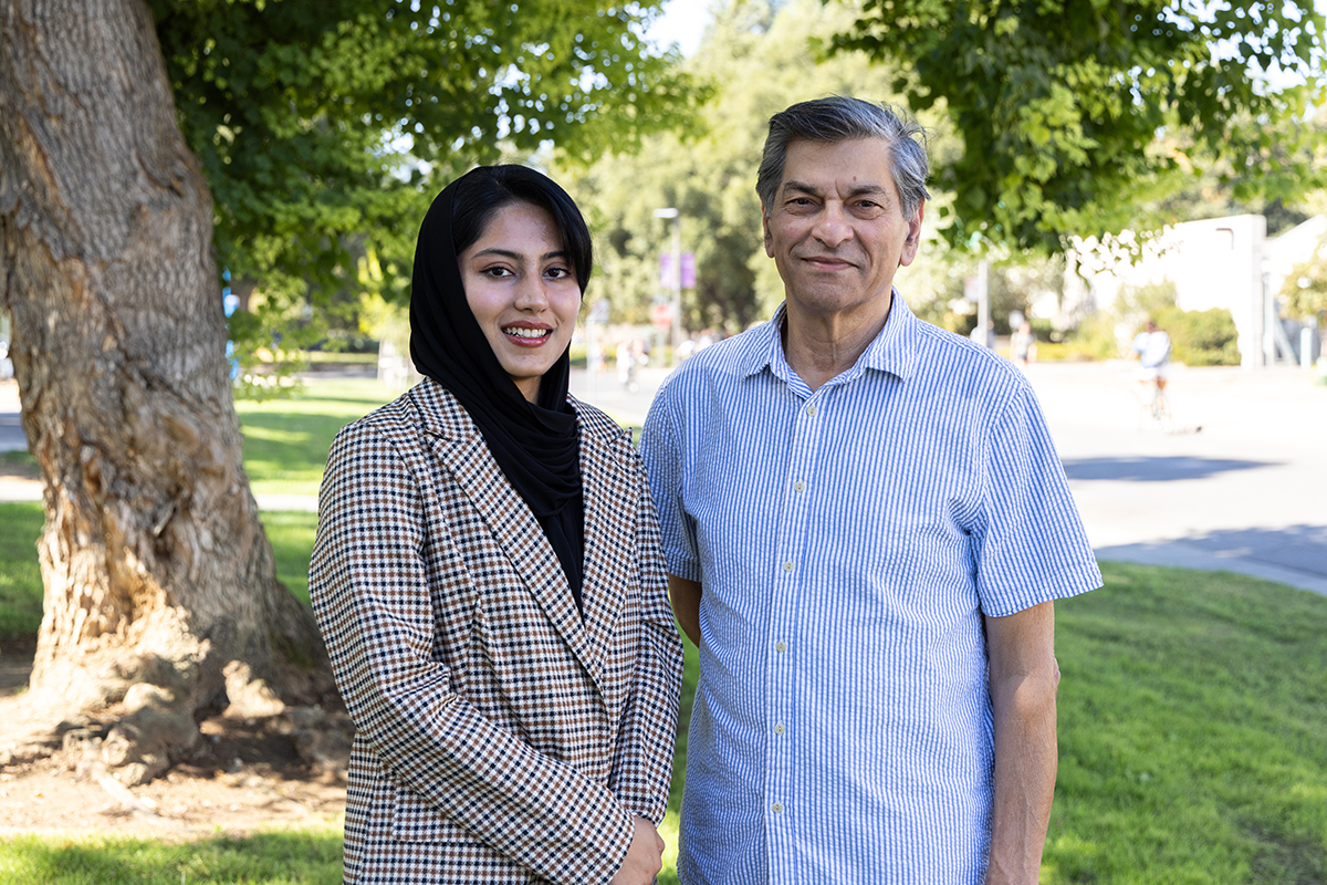 A young woman wearing a grey check jacket and black headscarf stands next to an older man wearing a light blue short-sleeved shirt. They are outdoors under a tree. 