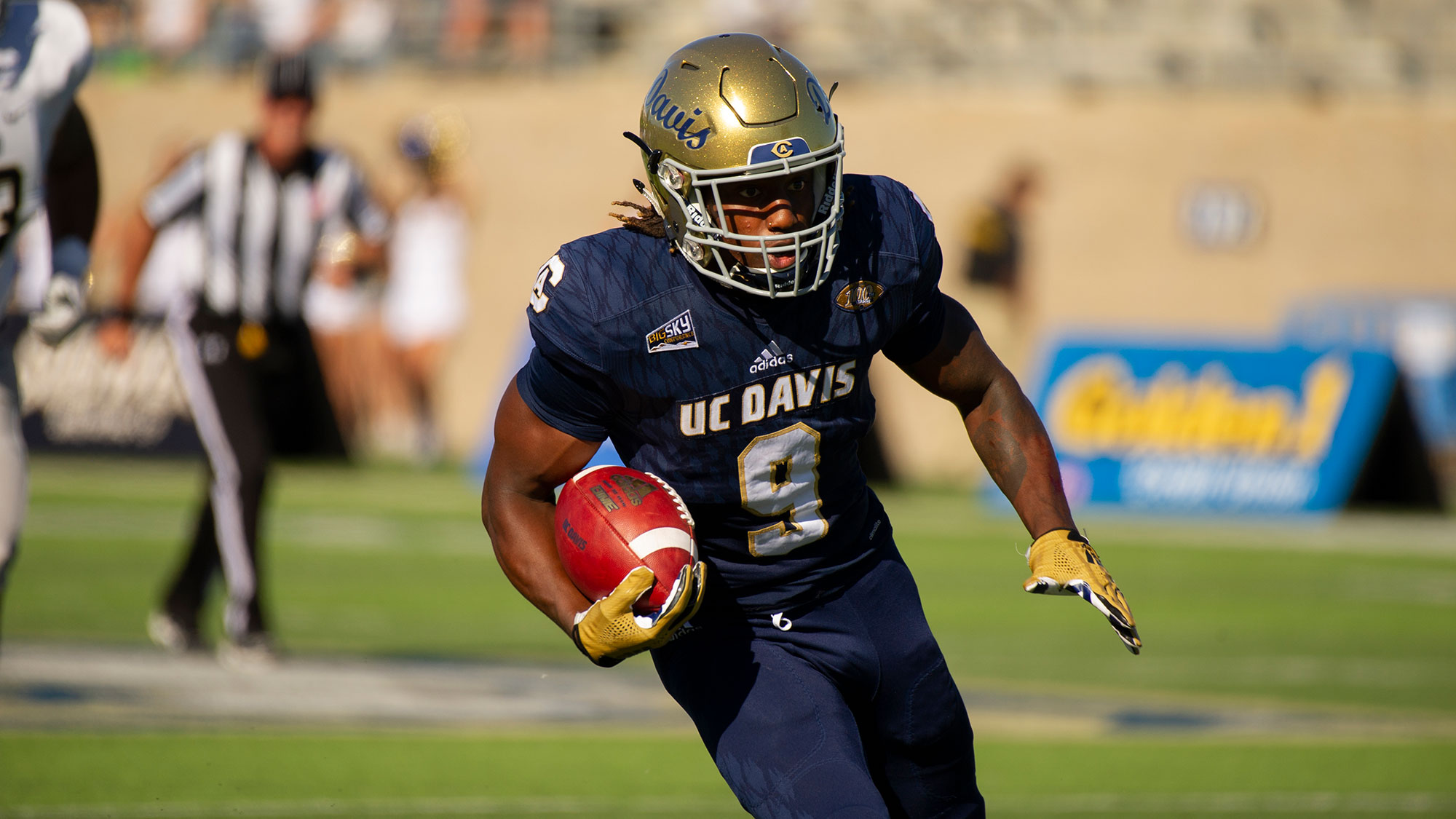 An Aggie football player runs with the ball during a game. A referee is seen running in the background.