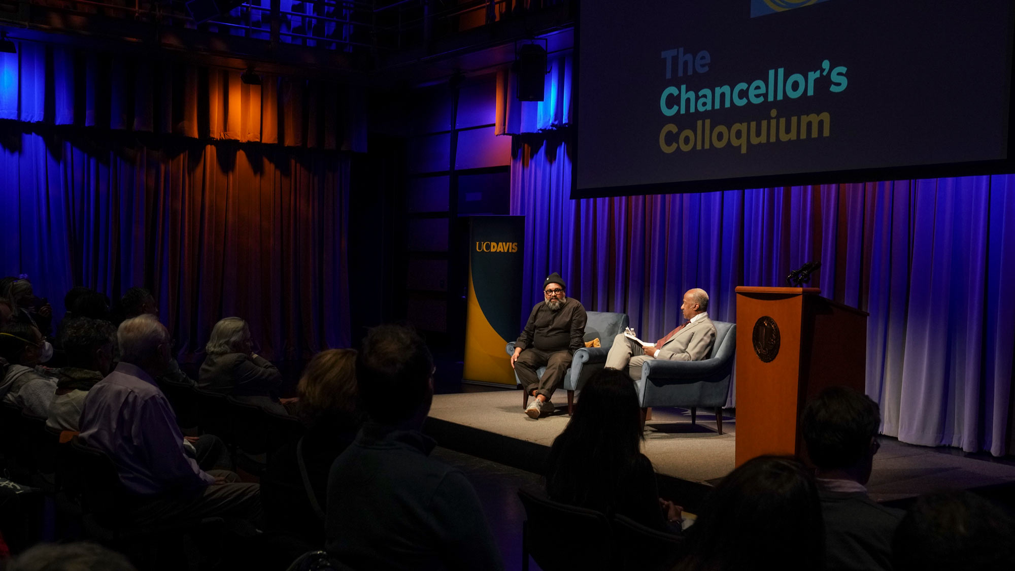 Joseph Patel and Chancellor May on stage, under "Chancellor's Colloquium banner; wide shot shows audience, too