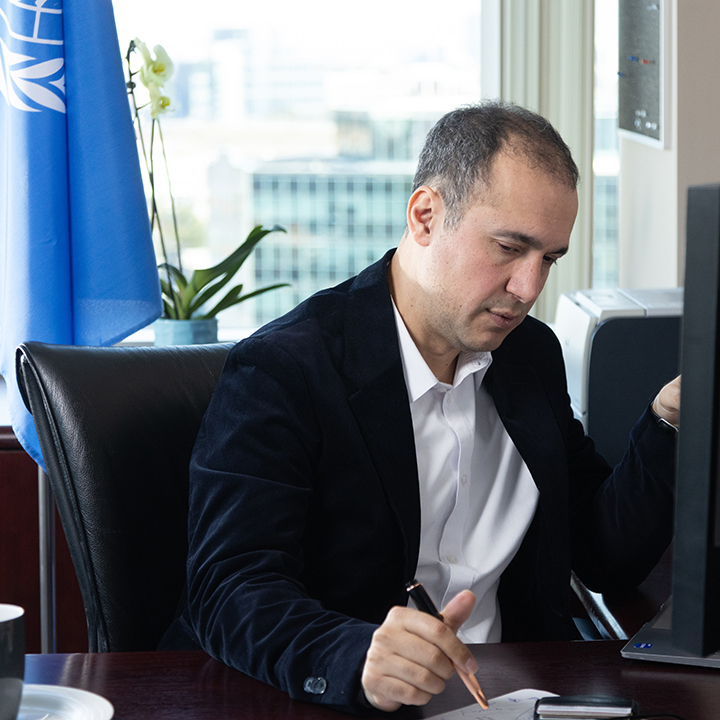 Man in blazer at office desk writing with pen, monitor and city view behind