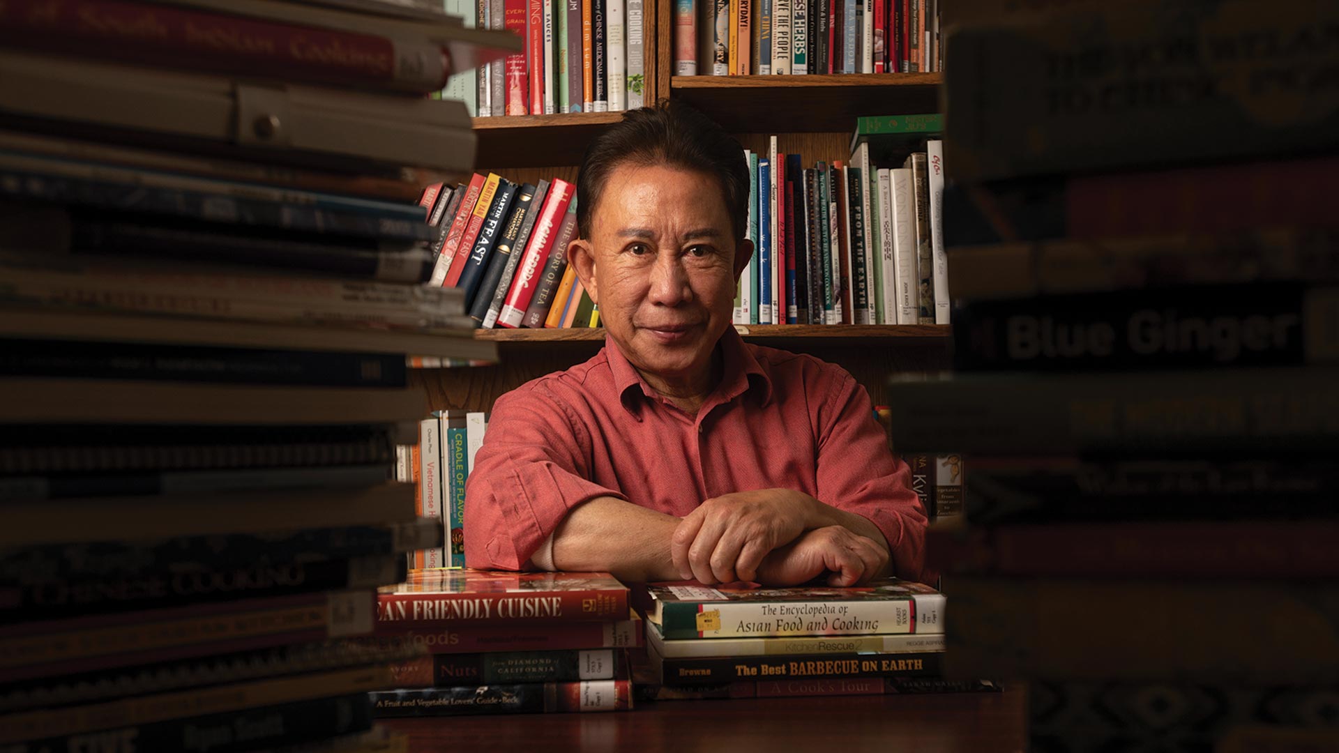 A man sits in front of a bookcase and is framed by books on the table in front of him. He looks directly into the camera.