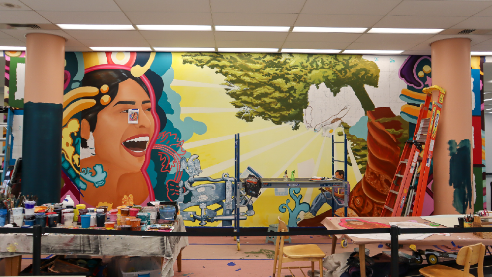 Bright mural in art studio: giant smiling face, tree, open book, scaffolding and paint supplies