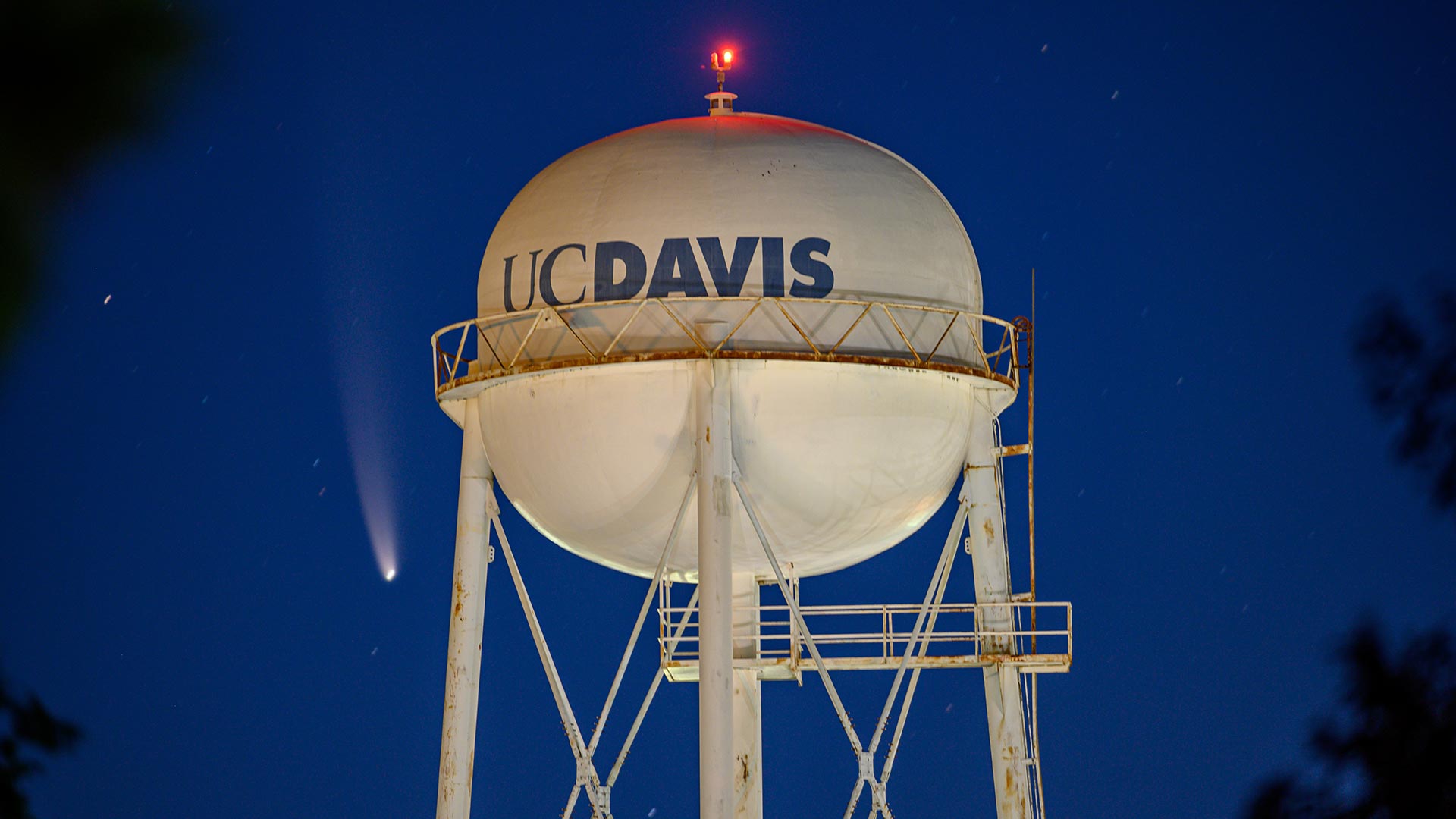 Night photo of a UC Davis water tower. Just to the left of the tower, a comet can be seen like a bright star, with a tail shining upwards from it.