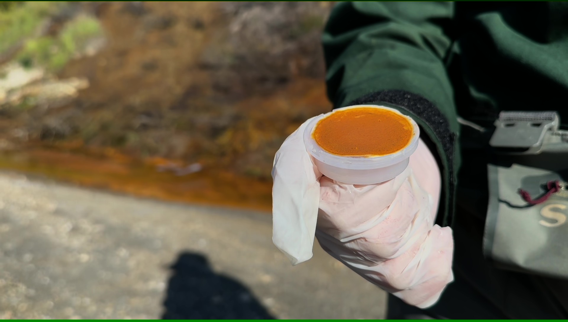 Gloved hand holding small white cup of orange substance beside rocky stream