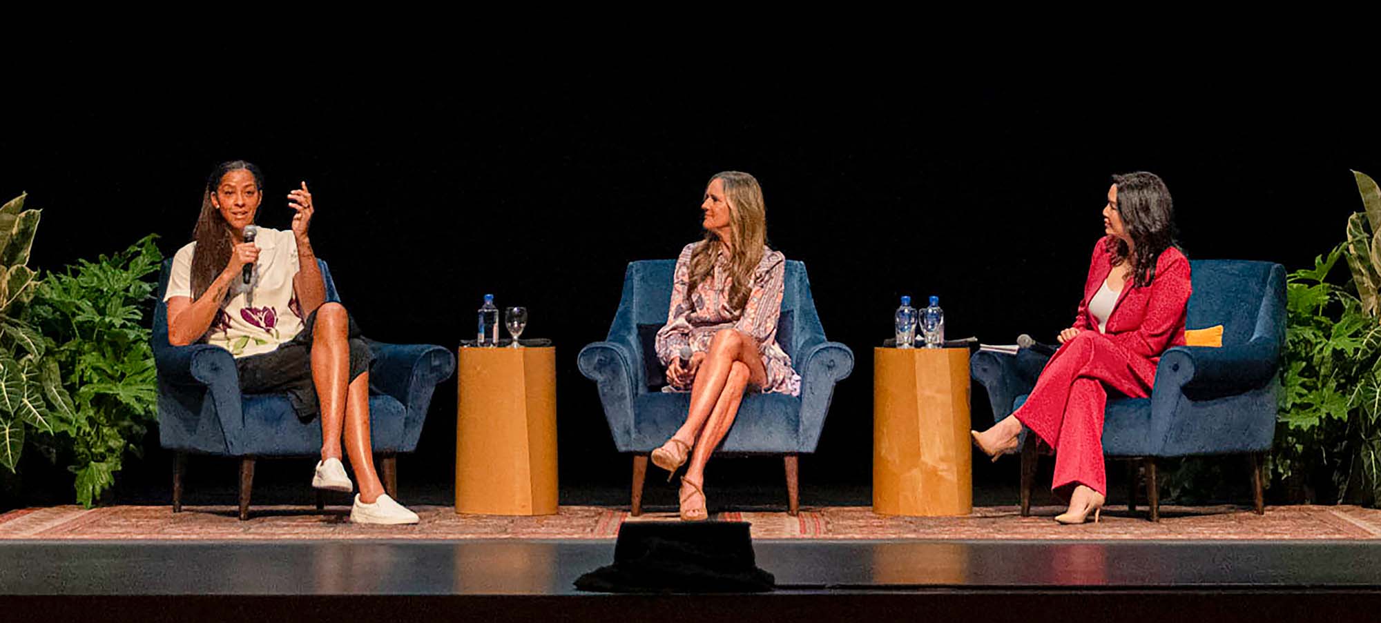 Three women seated on stage for a panel discussion, black backdrop and side plants.