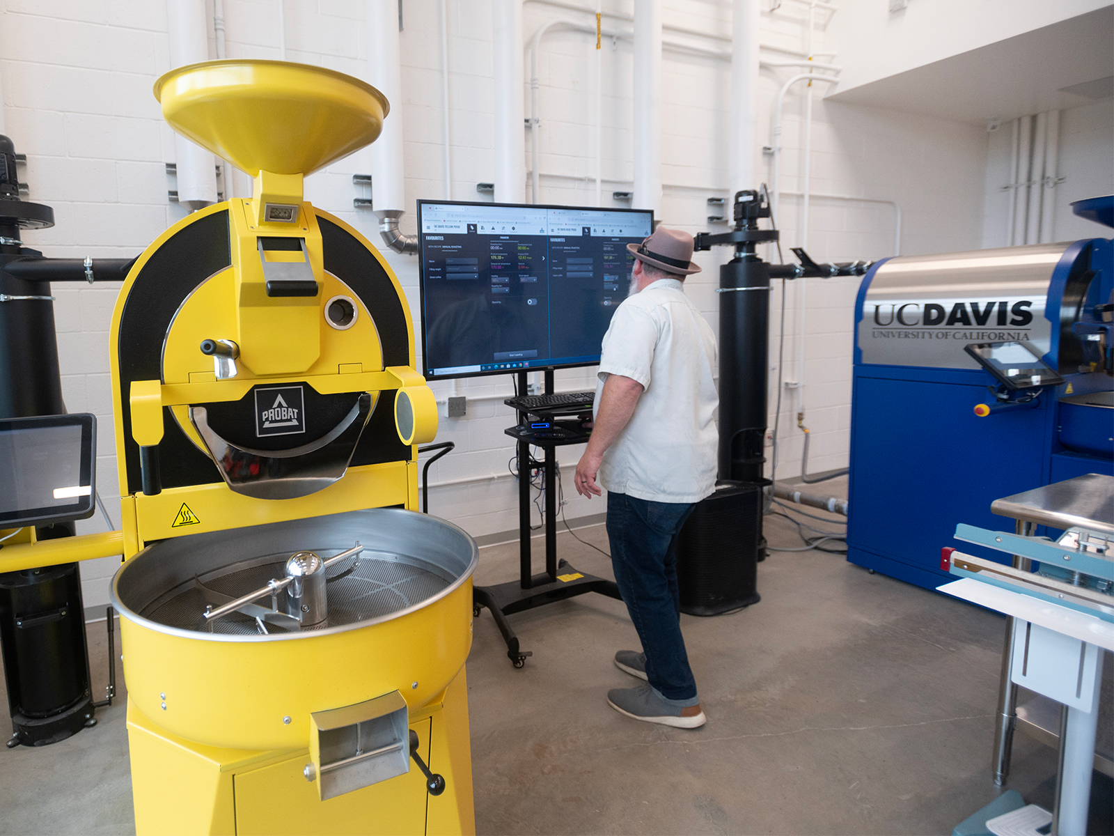 A man in a white, short-sleeved, button-up shirt, dark blue jeans, gray sneakers and a feathered gray fedora uses a computer with a large monitor between two large coffee roasters branded in UC Davis' Aggie Blue and Aggie Gold colors.