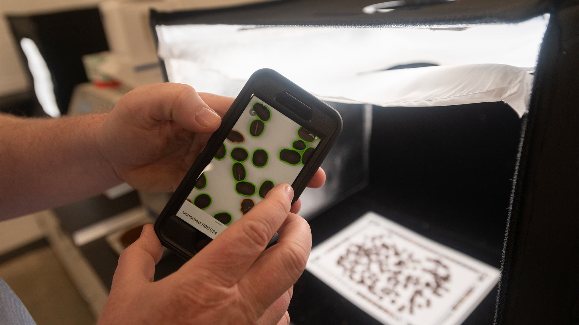 Coffee beans are strewn on a special paper for the Roastpic app. The beans are lit by a photo light box on a table. Professor Ristenpart reveals the photo on his phone he took using Roastpic, zooming in to see good-quality coffee beans outlined in green and a couple of beans outlined in orange.