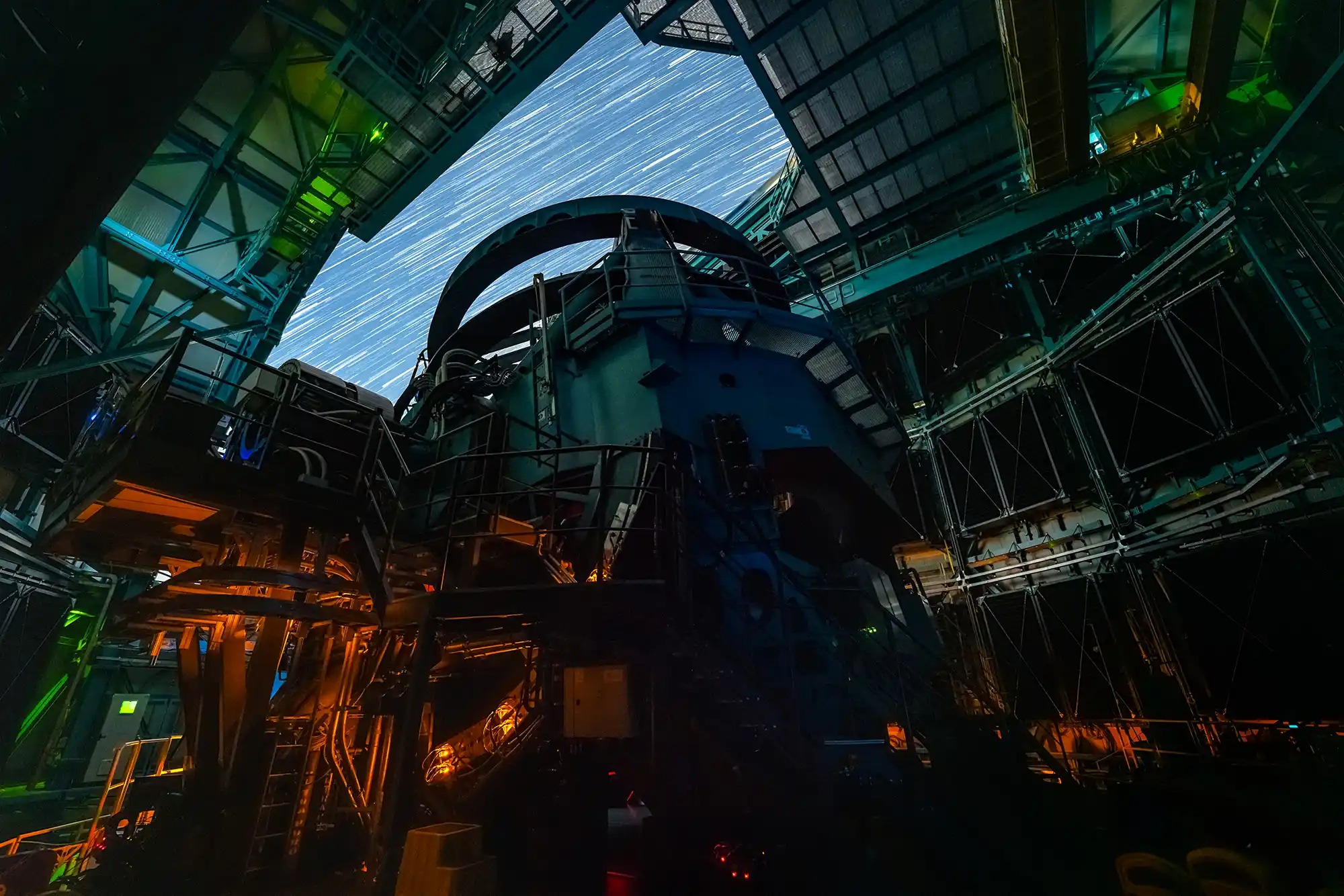 Large astronomical telescope inside open observatory dome, lit in green and orange