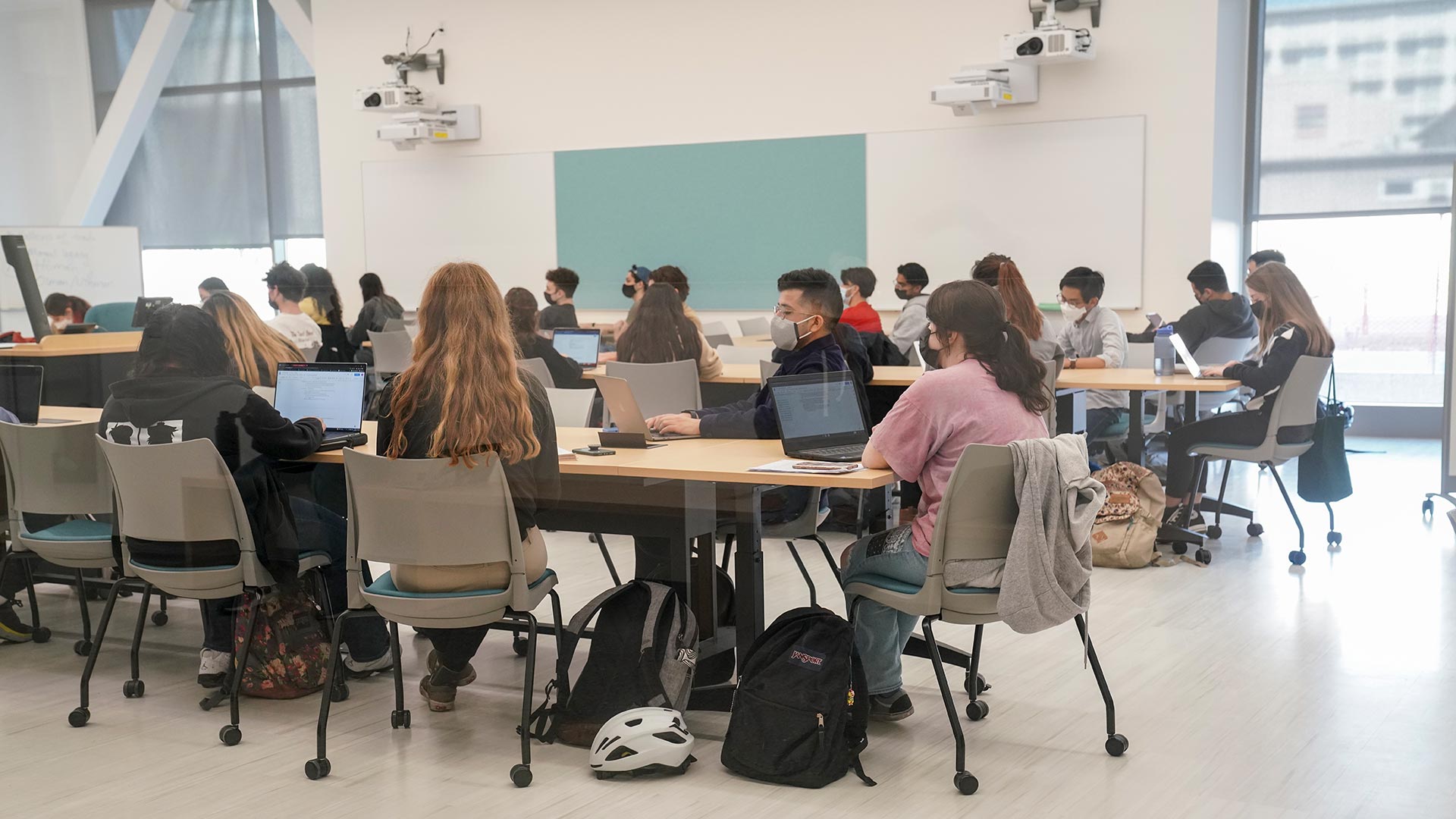 Students sit at tables with their laptops in a college classroom. There are large windows along one side, a blackboard and white boards on the wall, and audio/visual equipment in the ceiling.