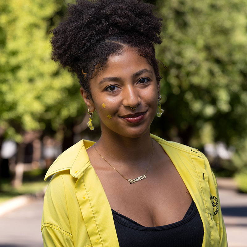 Portrait photo of a student wearing a black shirt with yellow collared short over it, smiling at the camera and has black hair pulled up over their head.