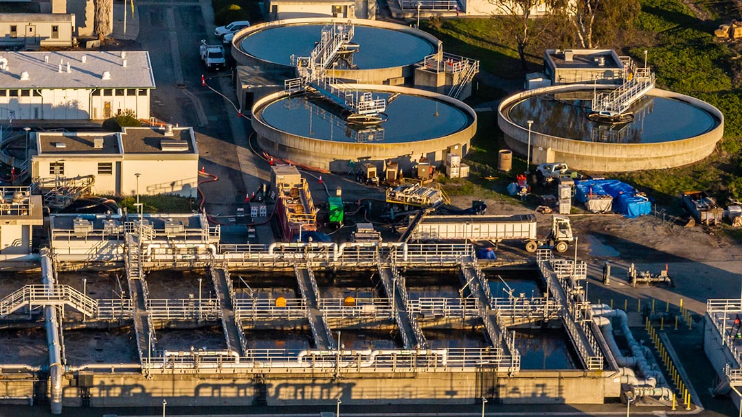 Aerial view of a wastewater treatment plant. Large round, open tanks are seen, as well as many pipes and small pools of water nearby.